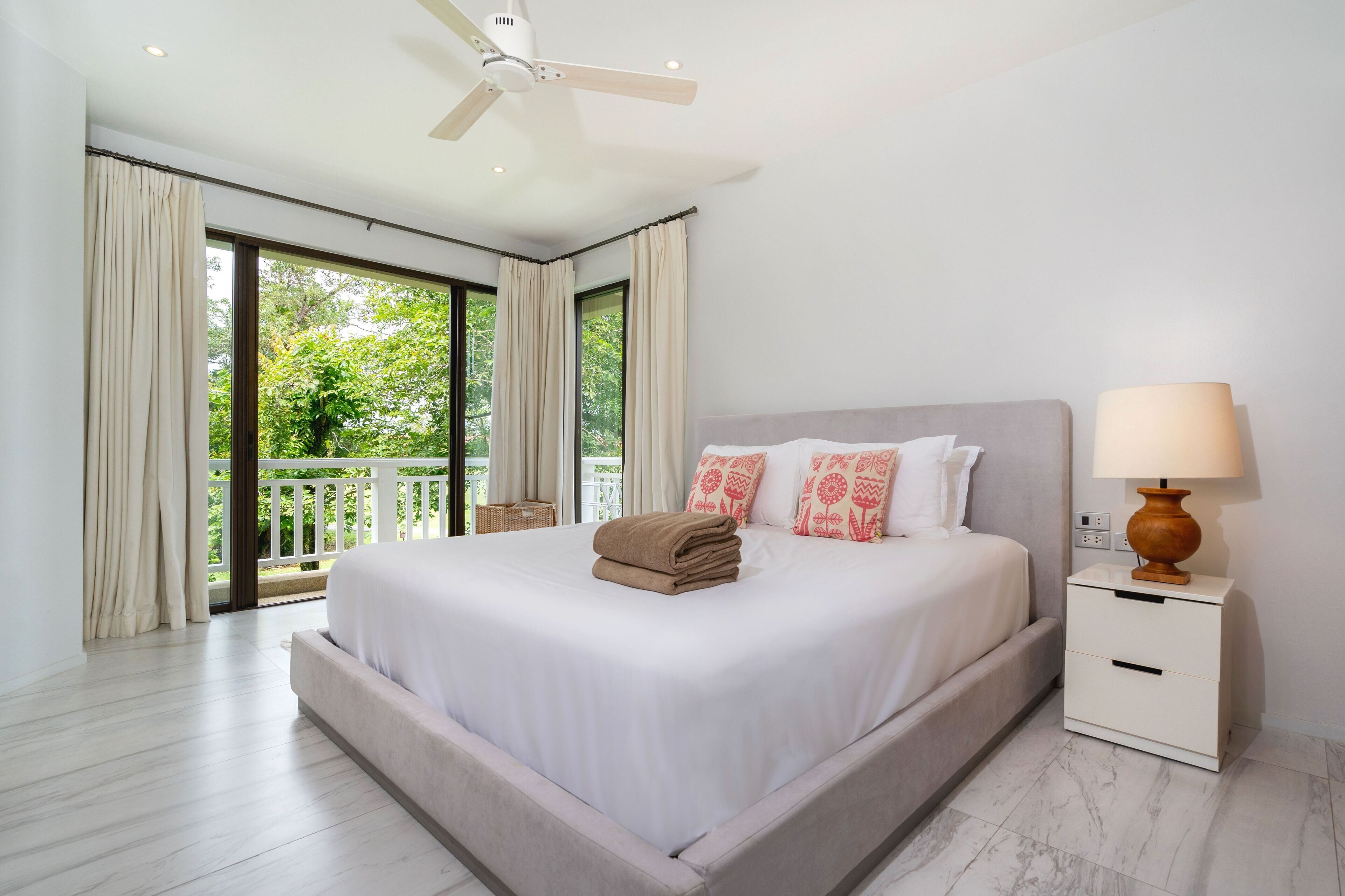 A bedroom with a large bed, a nightstand, a lamp, and a view of greenery through the window.