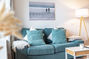 A teal sofa with pillows, a white lamp, and a framed picture of two people on a beach.