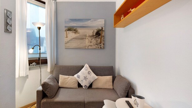 A living room with a sofa, a painting of a beach, a lamp, and a shelf with shoes. - Apartment 'Ferienwohnung Strandquartier' with Private Terrace and Wi-Fi (Lübeck Travemünde)