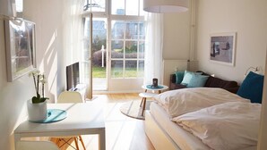 A bedroom with a bed, a desk, a chair, a table, a lamp, a radiator, and a window with curtains.