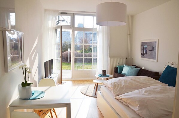 Une chambre à coucher comprenant un lit, un bureau, une chaise, une table, une lampe, un radiateur et une fenêtre avec des rideaux.