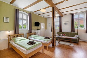 Una habitació amb dos llits de fusta, un sofà verd, una taula de fusta i grans finestres amb cortines.