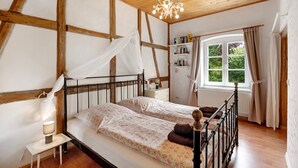 Un dormitorio con techo de madera, una cama con dosel, una mesita de noche con lámpara, una ventana con cortinas y una estantería.