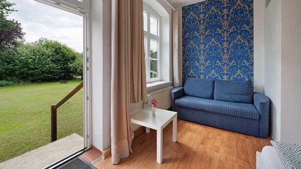 A room with a sofa, a small table, a window with curtains, and a patterned wall. - Room 'Blaues Zimmer' with Shared Garden and Wi-Fi (Schlagsdorf)