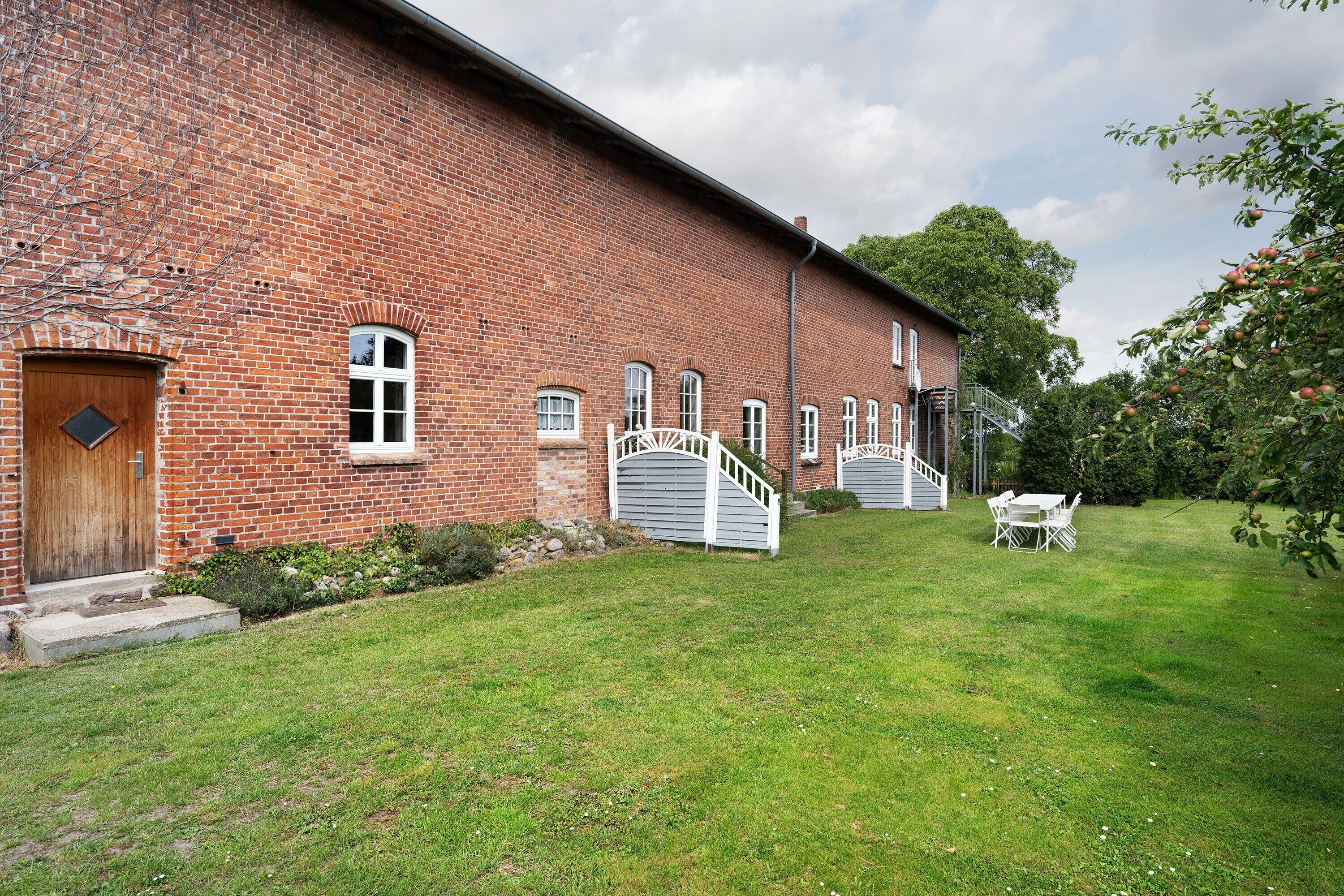 A brick building with a wooden door, white windows, and a white gate leading to a grassy area with a table and chairs.
