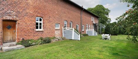 A brick building with a wooden door, white windows, and a white gate leading to a grassy area with a table and chairs.
