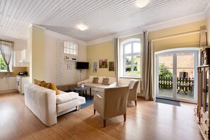 A living room with a sofa, armchair, and a television. There is a kitchen area with cabinets and a window.