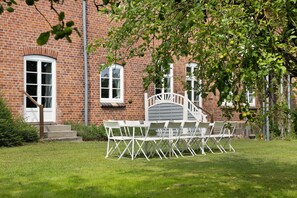 Un bâtiment en briques avec un escalier blanc, une pelouse et une table et des chaises en métal blanc disposées à l’extérieur.