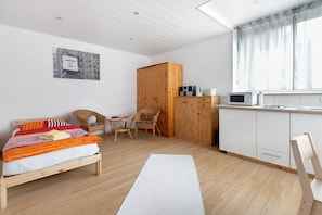 Una habitación con una cama, un armario de madera, una mesa de comedor con sillas, una zona de cocina con microondas y una ventana con cortinas.