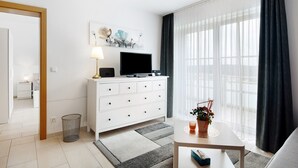 A modern hotel room with a white dresser, a flat-screen TV, a small table with a plant, and a view of the outdoors through sheer curtains.