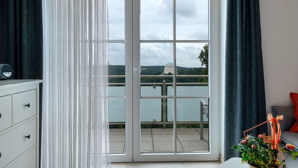 A room with a sliding glass door leading to a balcony with a view of a body of water and a distant building.