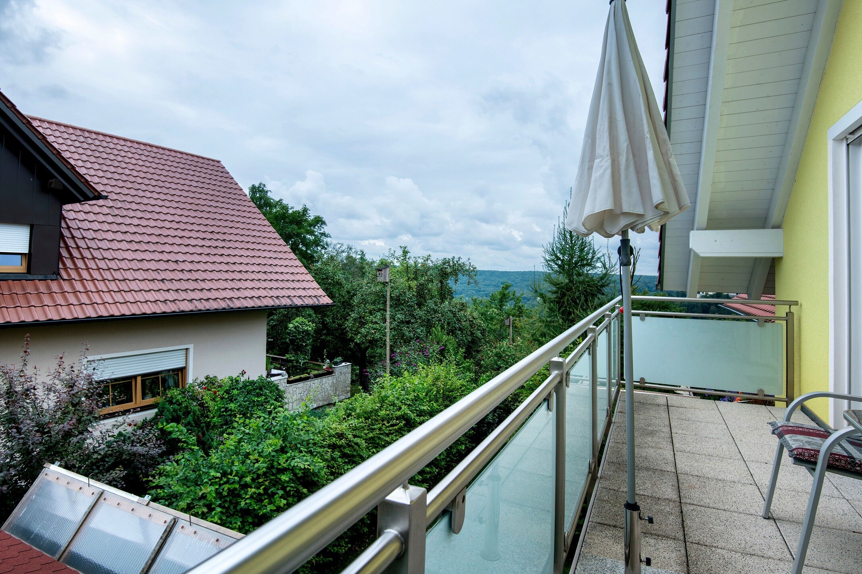 A balcony with a metal railing, a glass door, and a view of a red-roofed house and greenery.