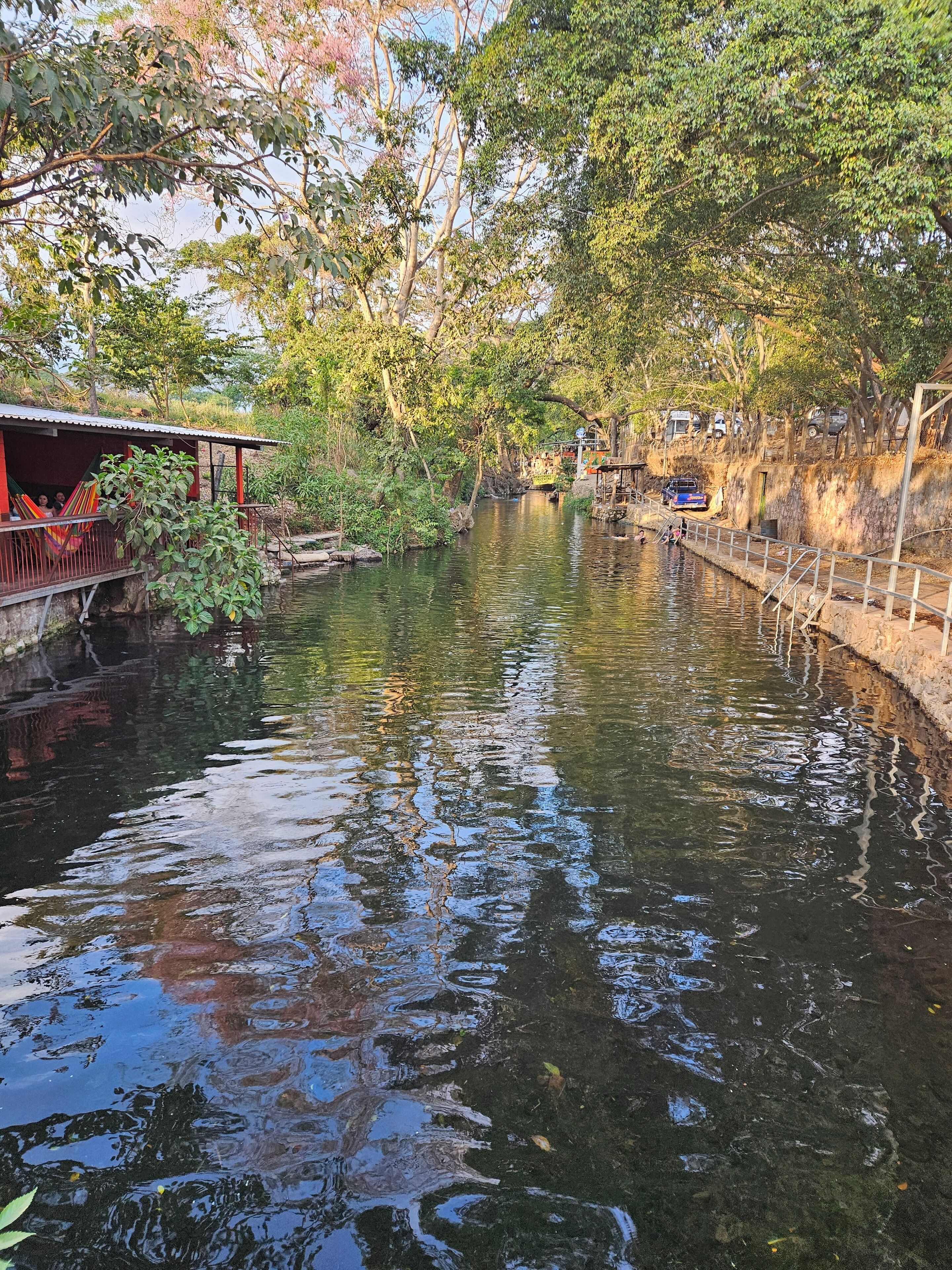 Blick auf einen Fluss mit einer Holzbrücke, einem kleinen Gebäude mit rotem Dach und einem in der Nähe geparkten blauen Fahrzeug.