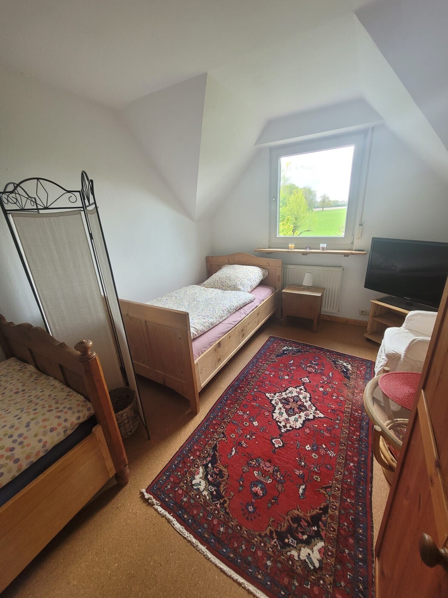 A bedroom with a bed, a window, a TV, and a red patterned rug.