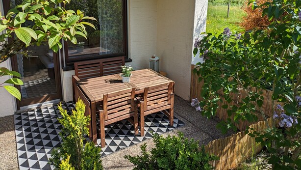 A patio with a wooden table and chairs, a checkered floor, and a view of a garden.