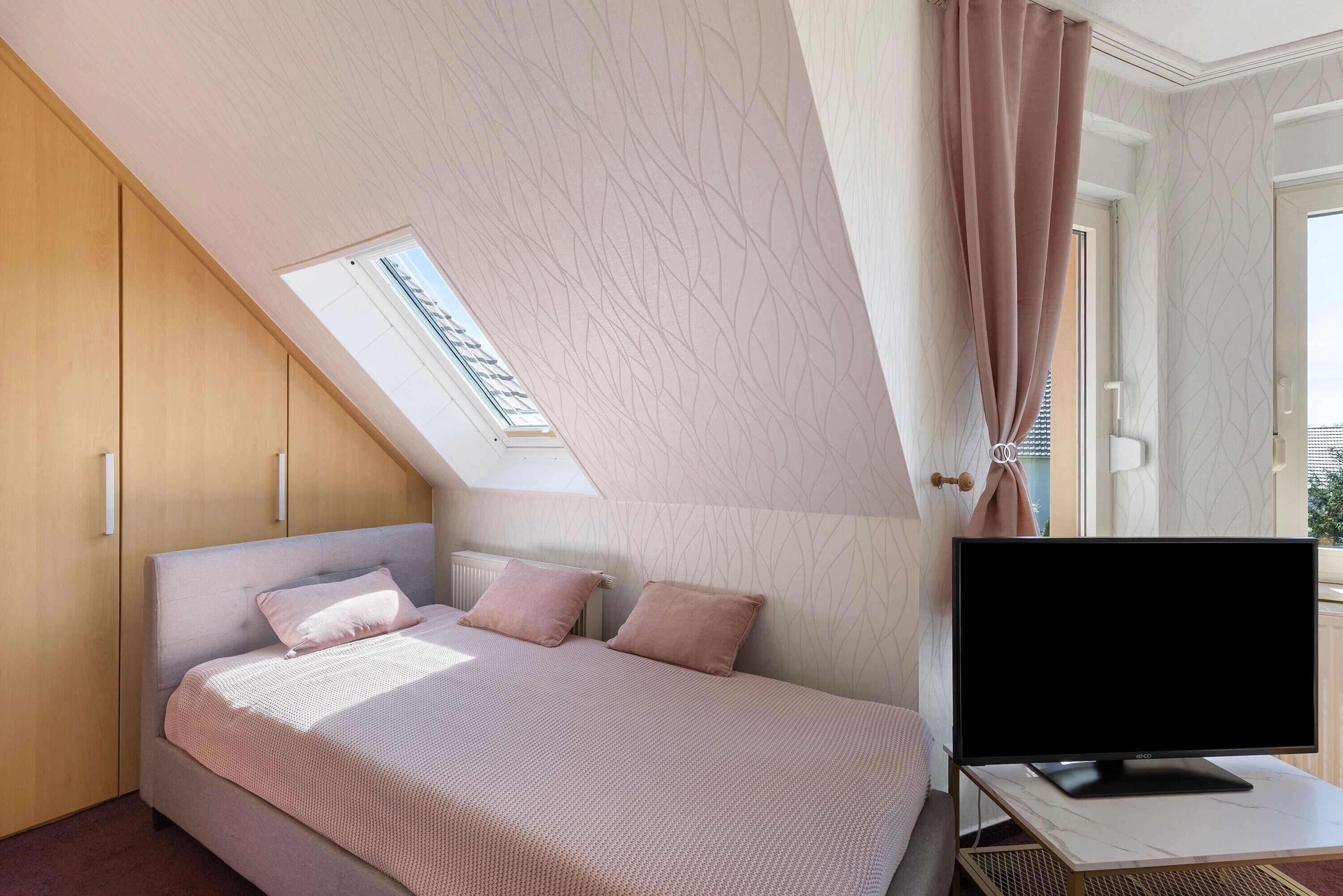 Een slaapkamer met een bed, een televisie op een standaard, een raam met gordijnen en een dakraam.