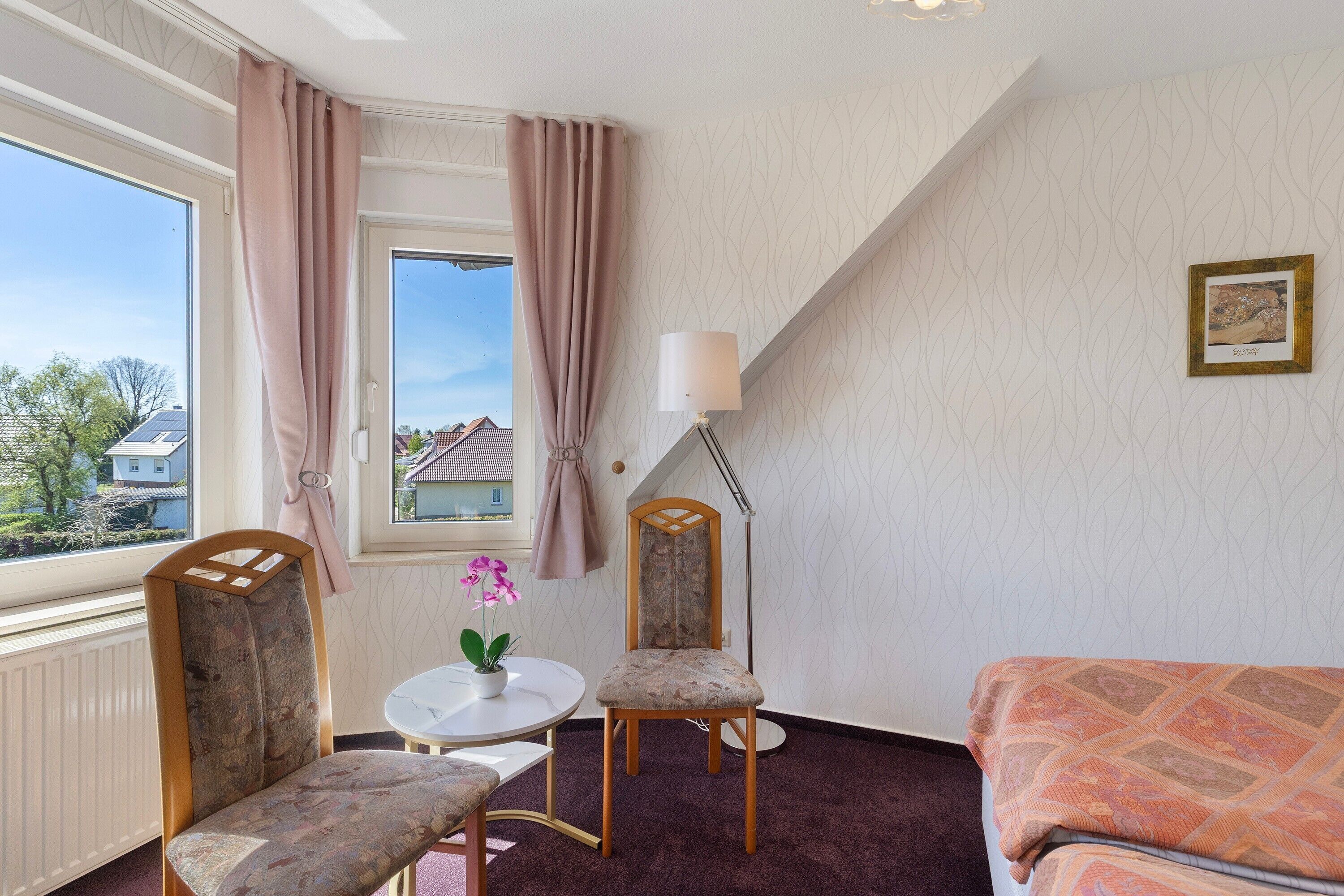 Ein Zimmer mit einem Bett, zwei Sesseln, einem Tisch mit einer Pflanze, einer Lampe und einem Fenster mit Vorhängen.