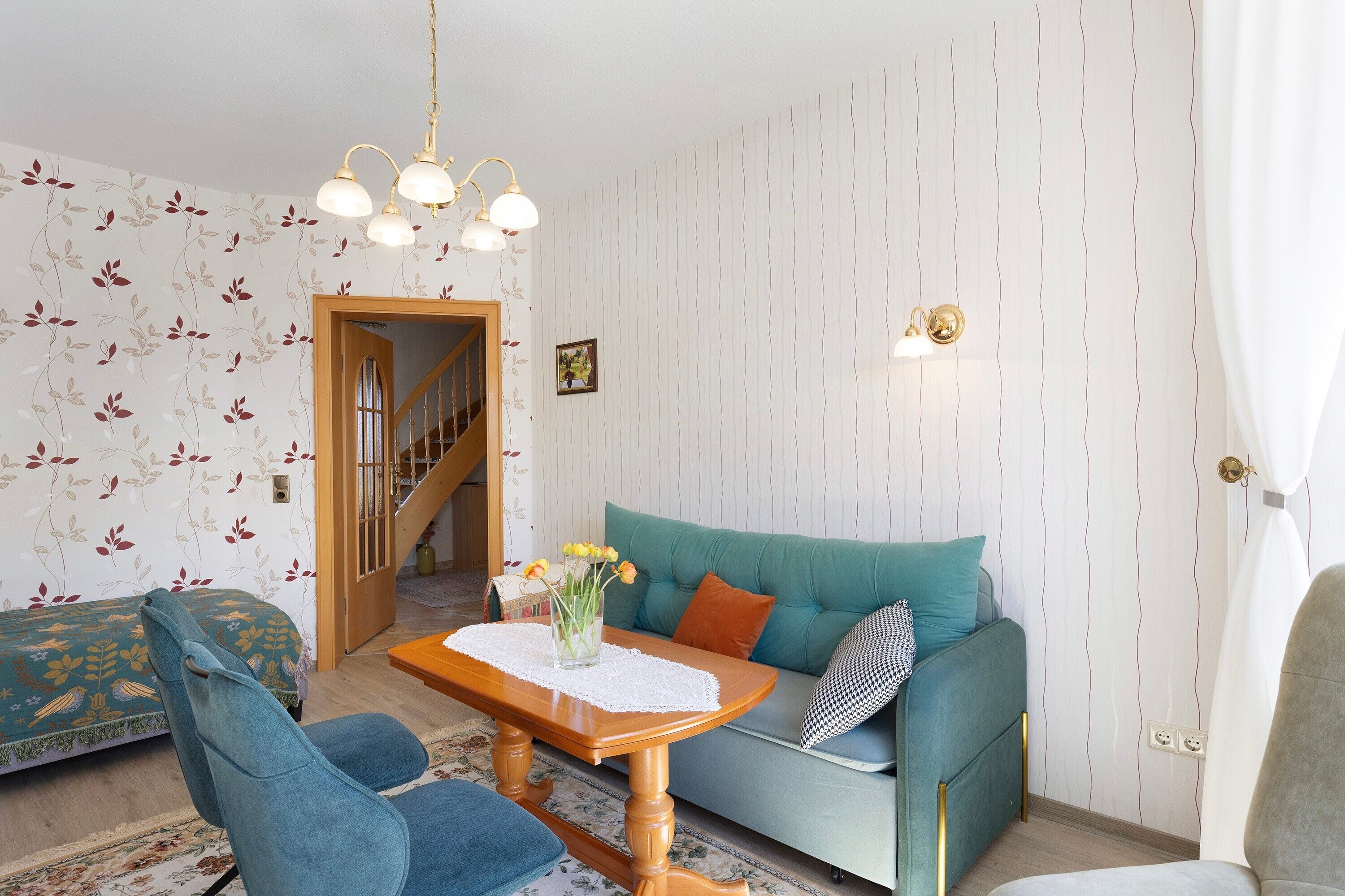 Ein Wohnzimmer mit Sofa, Sesseln, einem Tisch mit Blumenvase, einer Treppe und einer Wand mit vertikalen Linien.