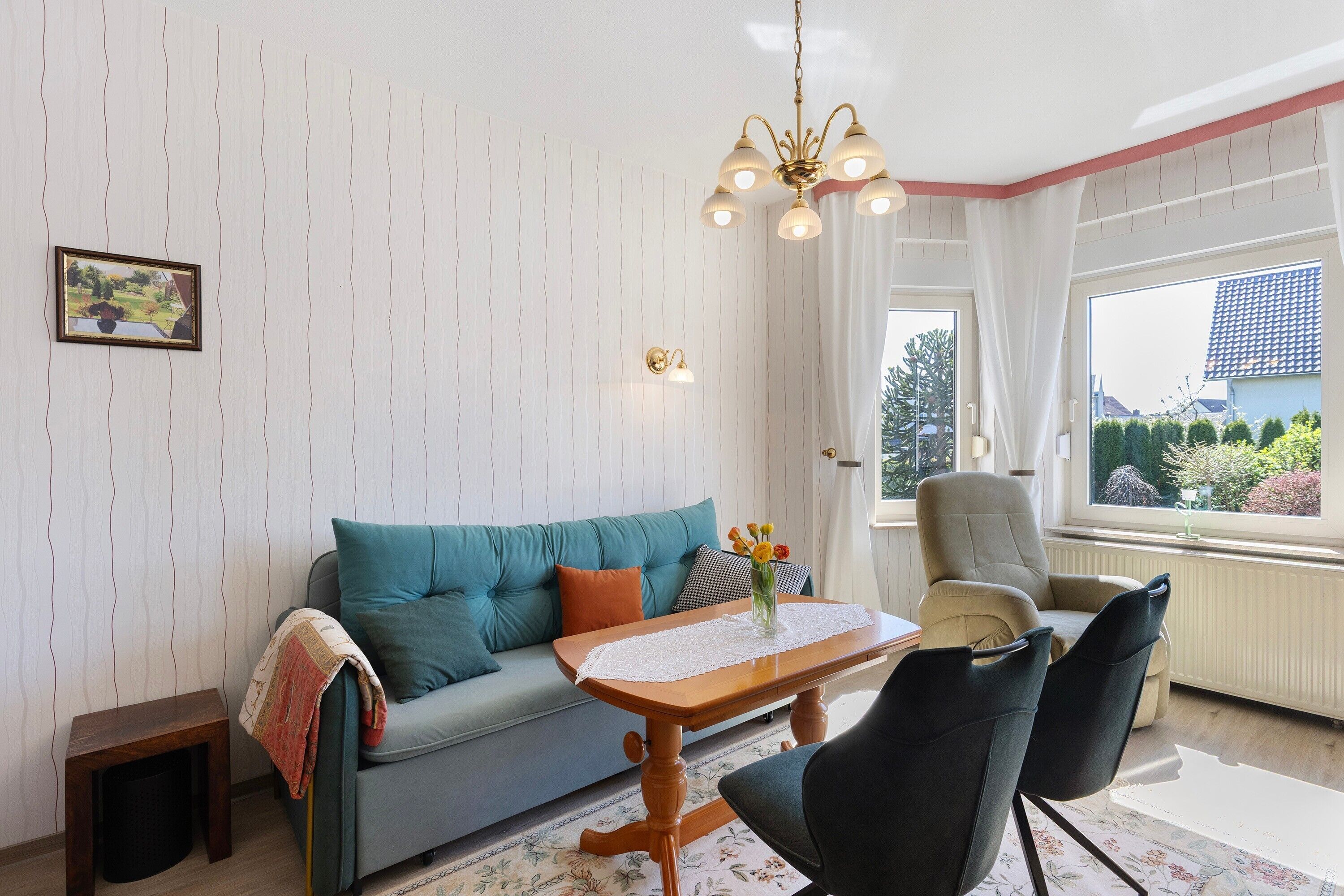 Ein Wohnzimmer mit einer Couch, einem Tisch mit Blumen in einer Vase, einem Sessel, einem kleinen Tisch und einem Bild an der Wand.