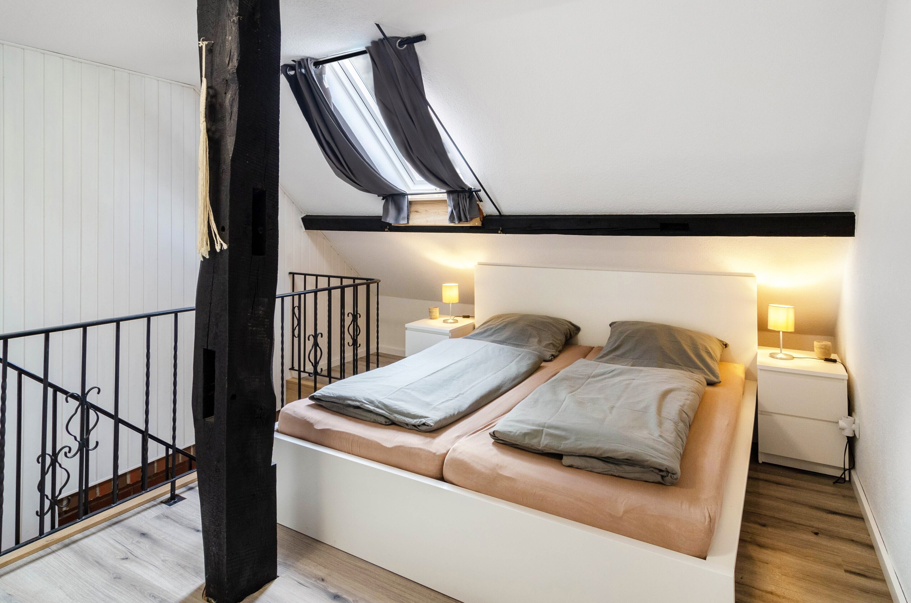 Quarto com cama, mesas de cabeceira, janela com cortinas, piso de madeira e escada.
