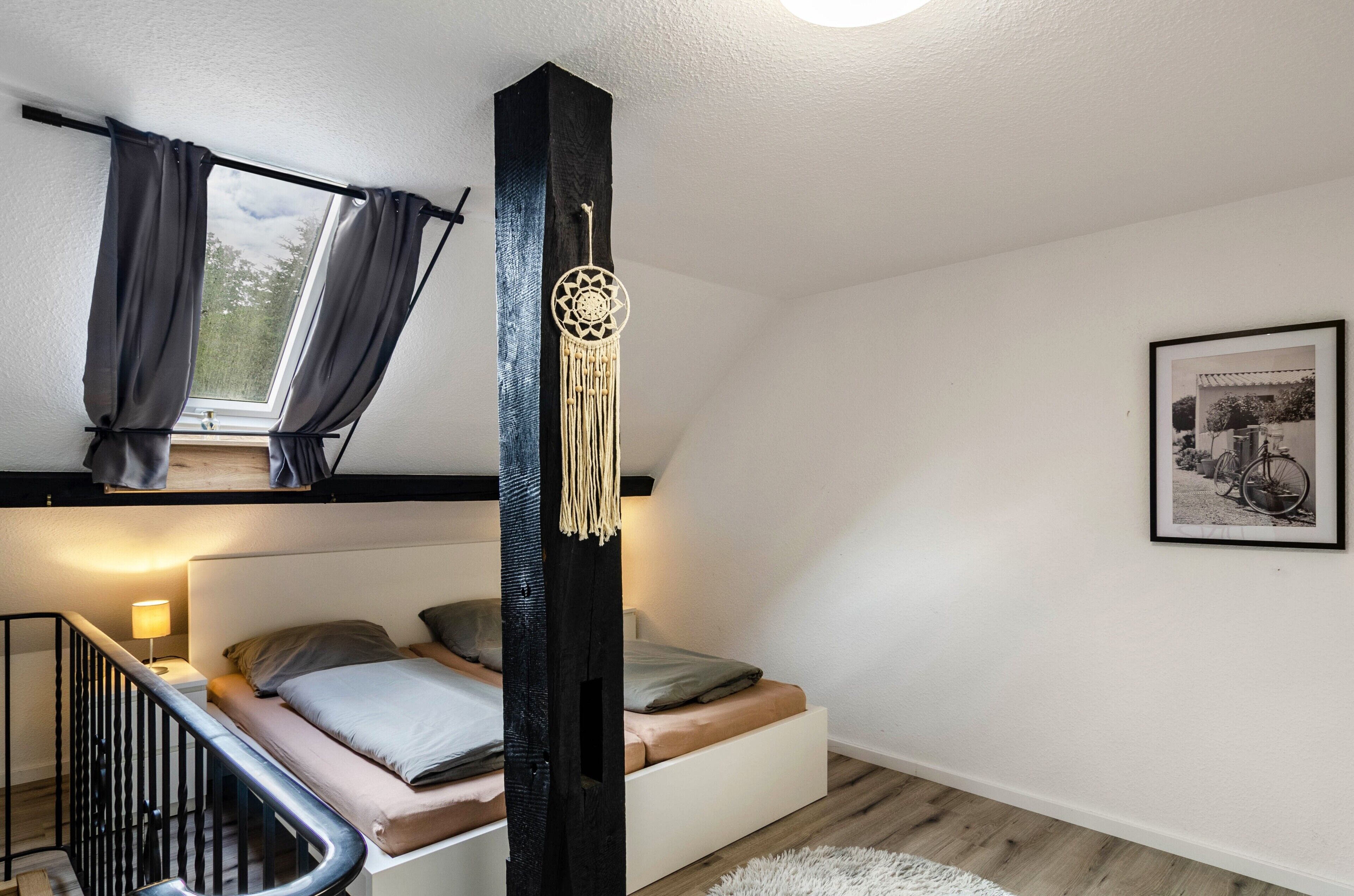 Une chambre avec un lit superposé, une fenêtre avec des rideaux, un attrape-rêves, un tableau encadré accroché au mur et un escalier.