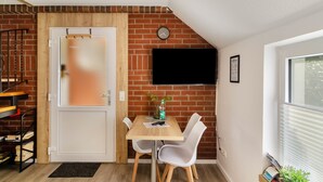 Una habitación con pared de ladrillo, puerta blanca, televisor montado en la pared, mesa de madera con sillas y ventana con persianas.