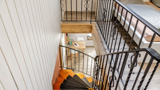 Una escalera con escalones de madera y barandillas negras que conduce a un dormitorio con cama y escritorio.