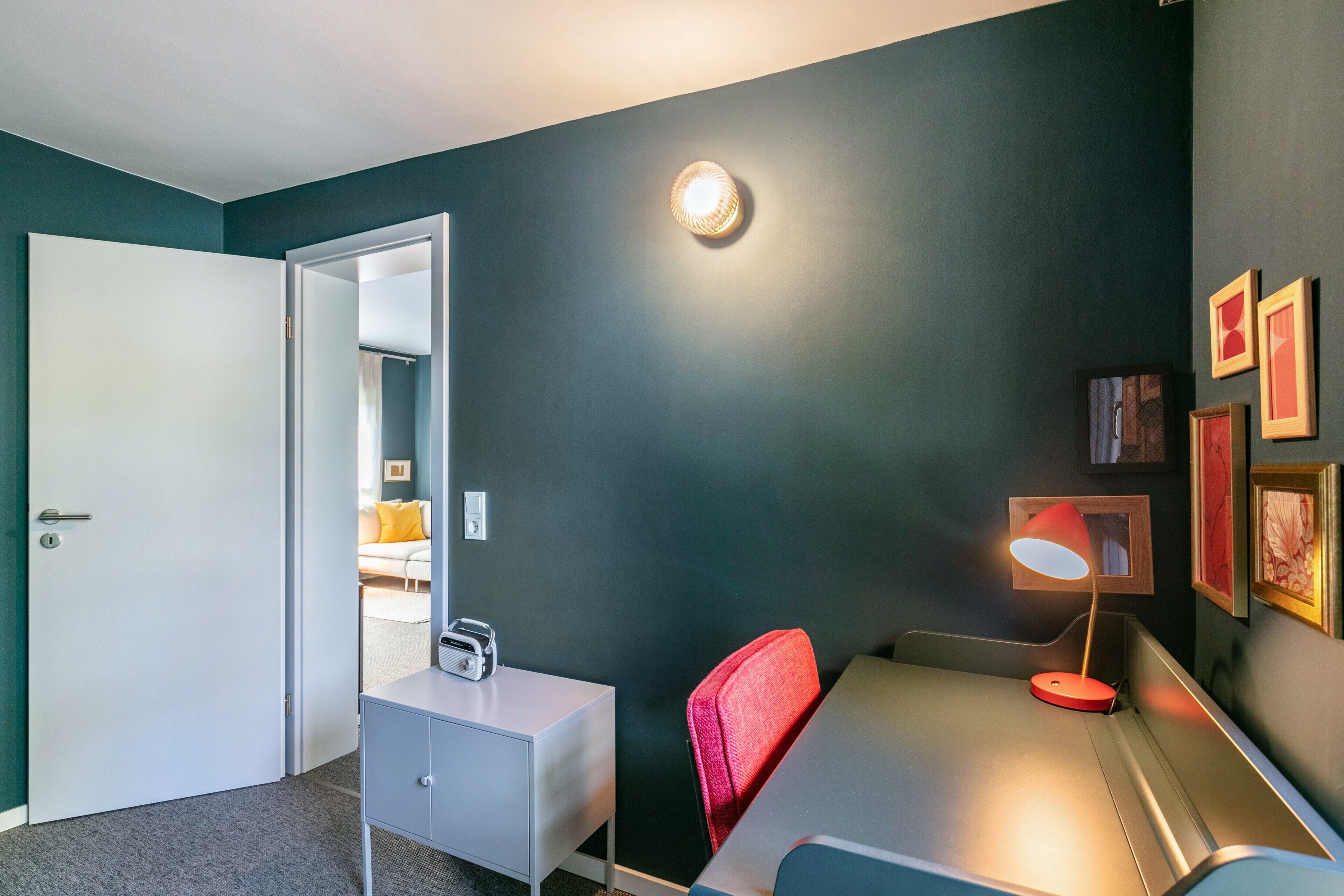 A room with a desk, chair, lamp, and a door leading to another room.