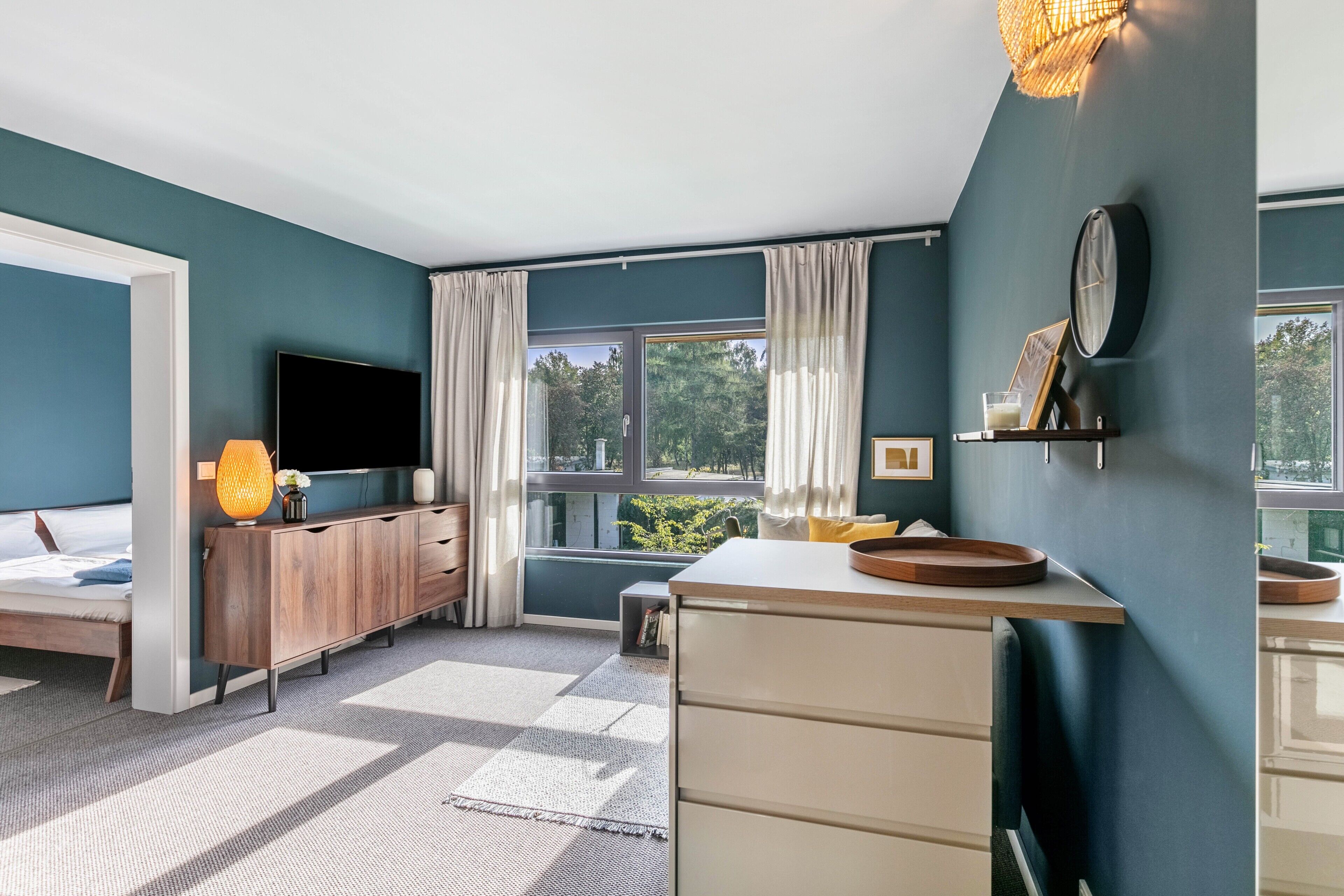 Un dormitorio moderno con cama, televisor, mueble de madera y un gran ventanal con cortinas.