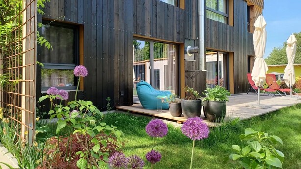Una casa moderna con fachada de madera, un patio con mobiliario exterior y un jardín con flores moradas.