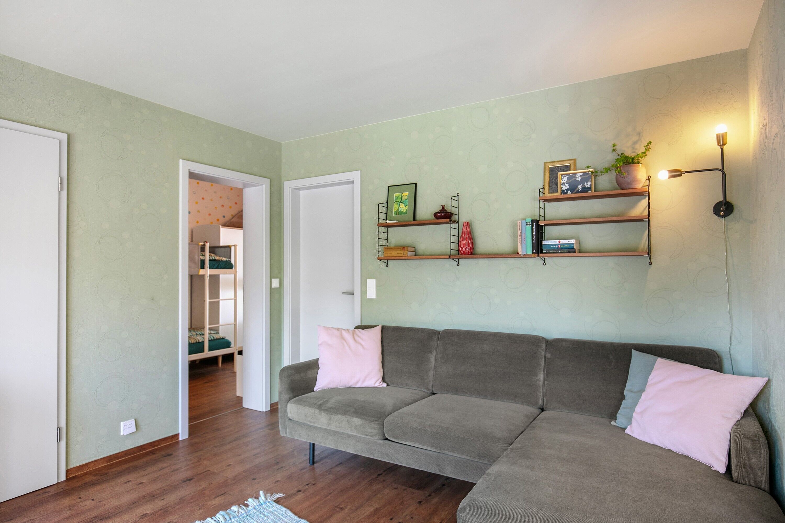 Un salón con un sofá gris, suelo de madera, una estantería empotrada con libros y objetos decorativos, y una puerta que da a otra habitación.