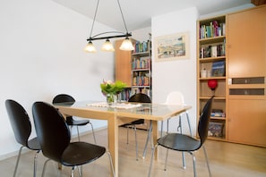 A dining area with a wooden table, black chairs, a vase with flowers, and a bookshelf with books.