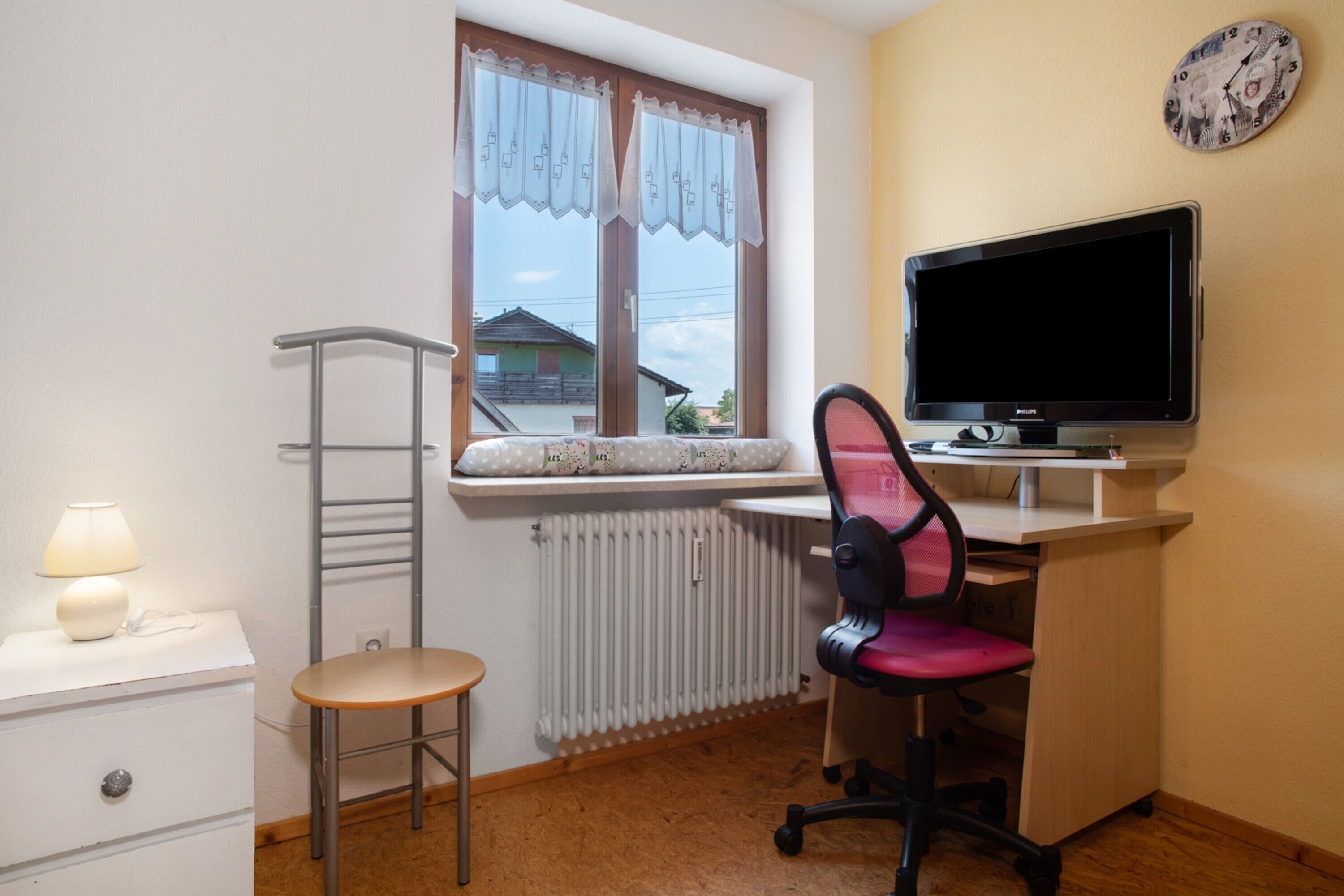 Ein kleiner Raum mit Schreibtisch, Stuhl, Heizkörper, Fenster mit Vorhängen und einer Uhr an der Wand.