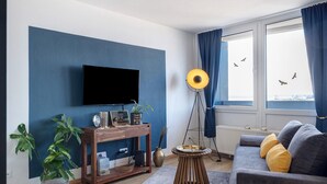 A living room with a flat-screen TV, a wooden side table, a floor lamp, a sofa with cushions, and a large window with curtains. - Apartment 'Clara' with Sea View, Balcony and Wi-Fi (Bremerhaven)