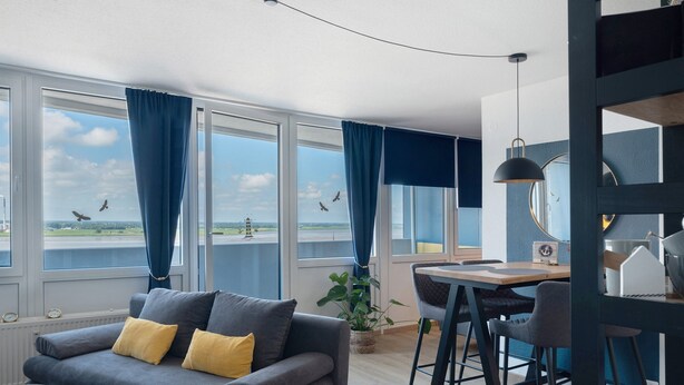 A modern room with a sofa, dining table, and large windows with curtains. - Apartment 'Clara' with Sea View, Balcony and Wi-Fi (Bremerhaven)