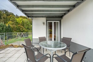 Une terrasse extérieure couverte, avec une table ronde en verre et des chaises en métal, un mur blanc et une rambarde en métal.