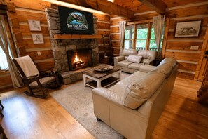 A log cabin interior with a stone fireplace, leather furniture, and a television displaying 'The Vacation Company' logo.