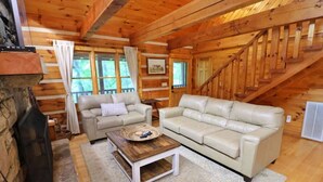 Ein gemütliches Cabin-Interieur mit Kamin aus Stein, zwei beigen Ledersofas, einem Holztisch und einer Treppe, die zu einer oberen Ebene führt.