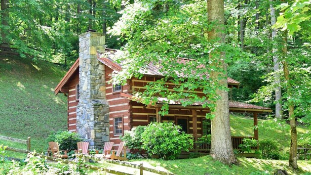 A log cabin with a stone chimney, surrounded by lush greenery and a wooden deck.