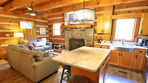 A cozy cabin interior with a stone fireplace, wooden walls, a dining area, and a kitchen with wooden cabinets.