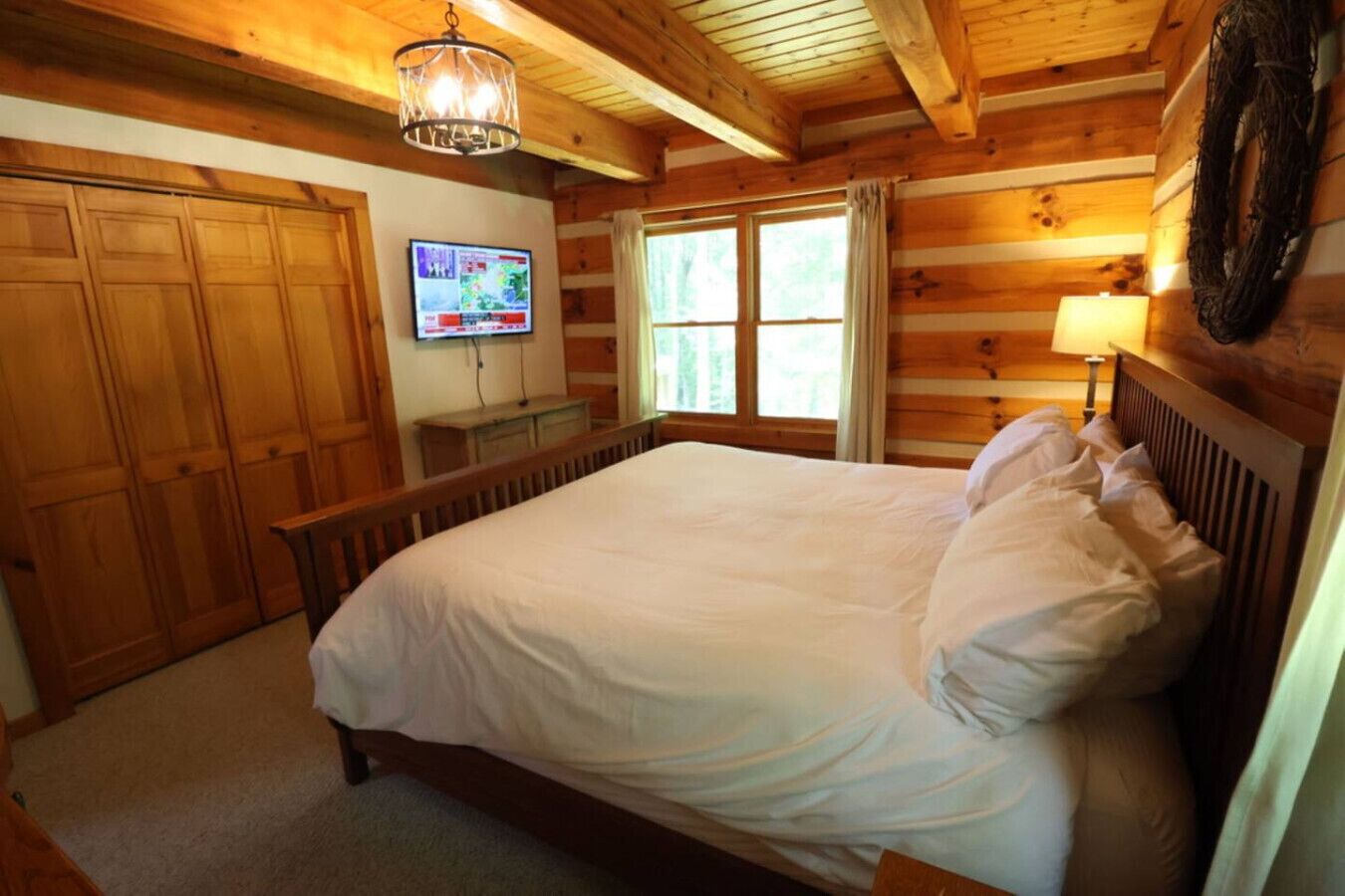 A wooden cabin bedroom with a bed, a television on the wall, and a window with curtains.