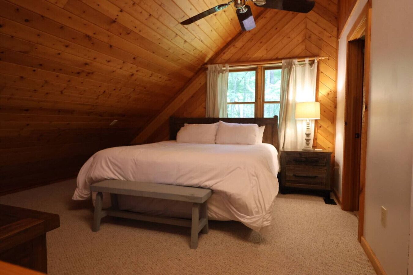 A cozy attic bedroom with a wooden sloped ceiling, a single bed with a bench, a nightstand, and a window with curtains.