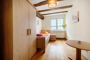 A bedroom with a bed, a bedside table, a wooden wardrobe, a window with sunflowers, and a small table with a bowl.