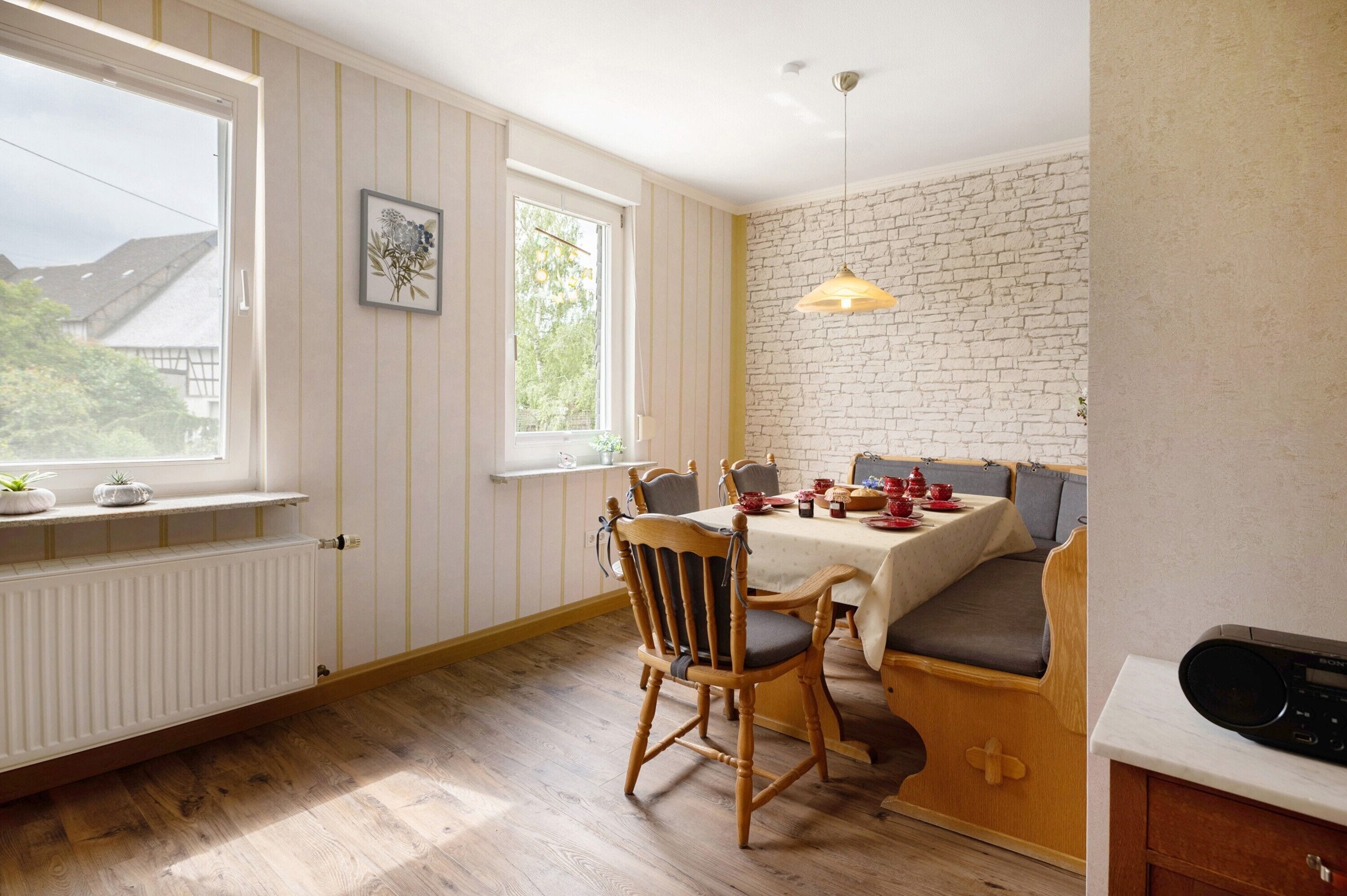 Un coin repas avec une table dressée pour quatre personnes, une chaise en bois, un banc, un radiateur et une fenêtre donnant sur des maisons.