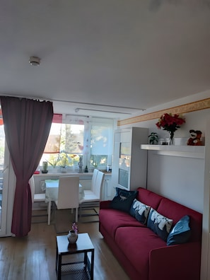 A living room with a red sofa, a small table, a white chair, and a window with curtains.