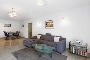 Una sala de estar con un sofá modular, una mesa de centro de vidrio, un comedor con sillas, una estantería e iluminación empotrada.