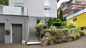 Un edificio de fachada gris, con una escalera que lleva a una puerta y un pequeño jardín con rocas y plantas.