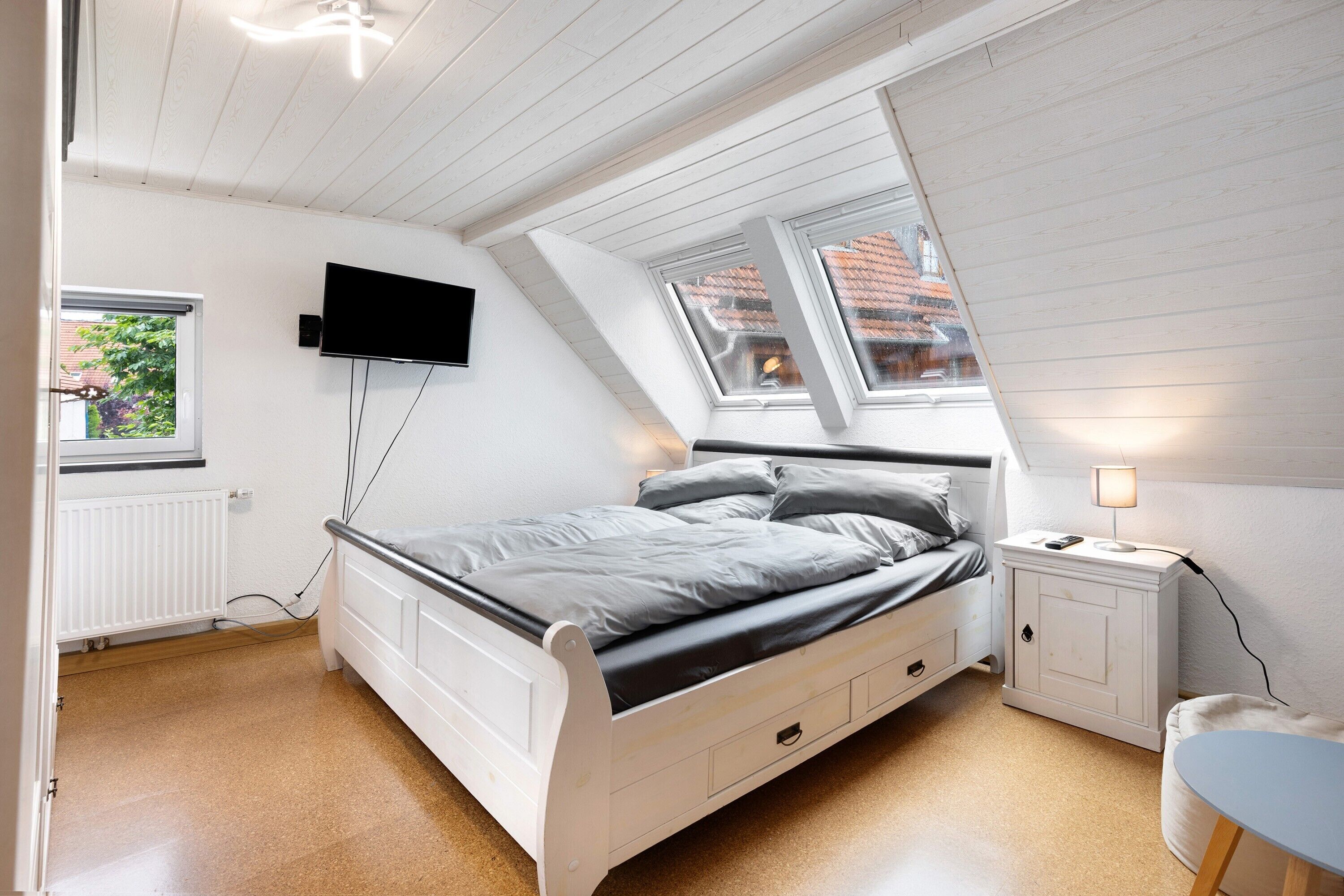 A bedroom with a bed, a TV mounted on the wall, a window, and a bedside table with a lamp.
