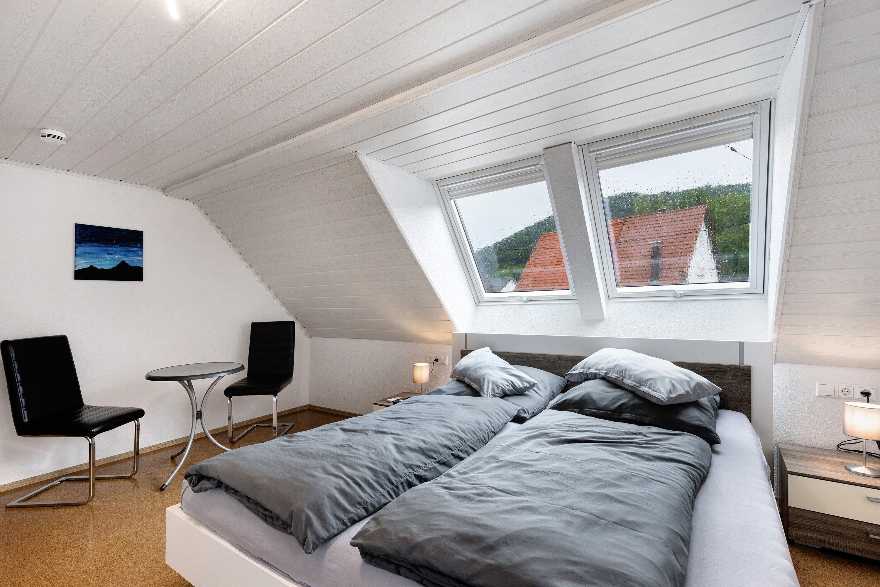 A bedroom with a bed, two chairs, a small table, a nightstand, and a window with a view of houses.