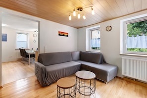 Un salon moderne avec un canapé d’angle gris, deux petites tables rondes, un coin repas avec une table et des chaises, et une grande fenêtre donnant sur un paysage verdoyant.
